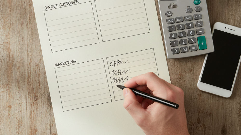 A hand writing on a one-page business plan template with sections for target customer, marketing, and financials visible on a wooden desk.