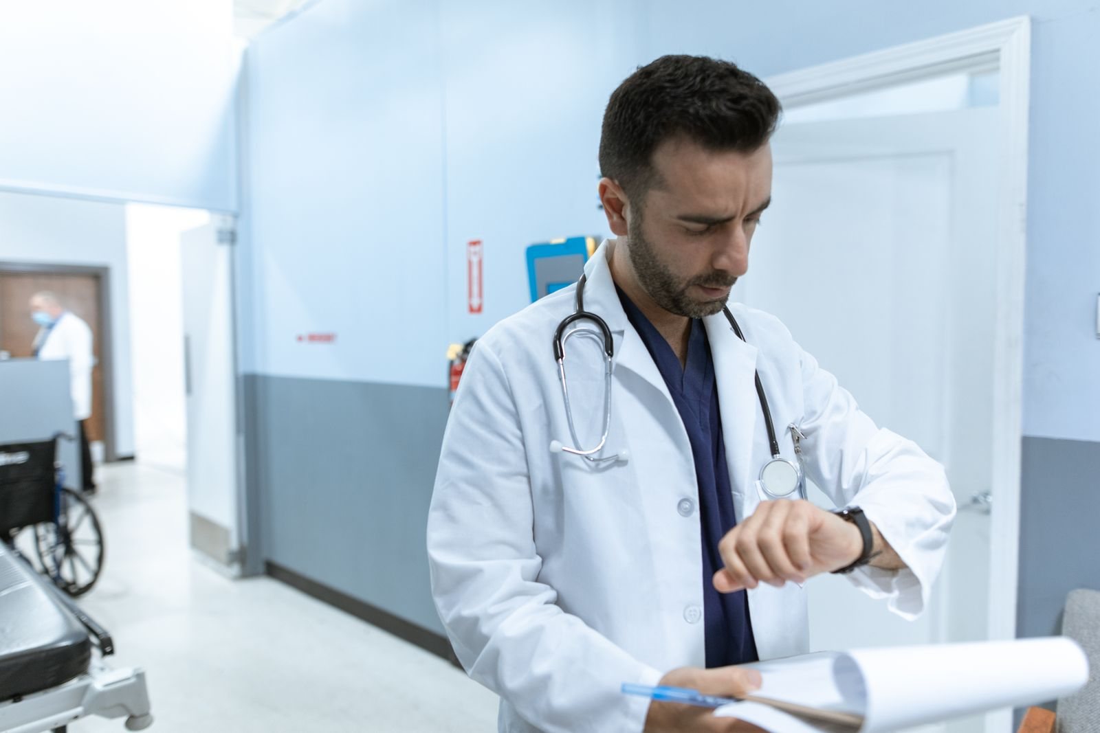 A doctor checking his watch in a hospital hallway, chart in hand