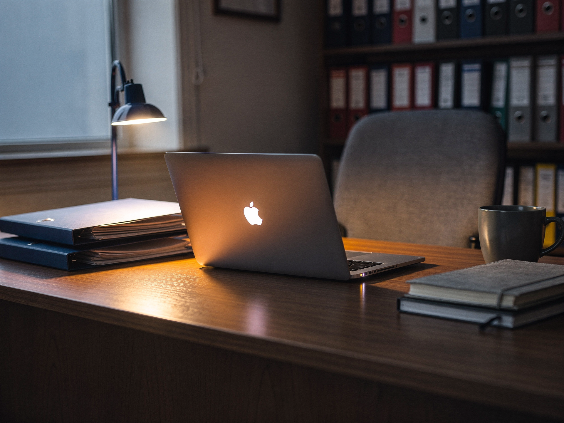 A laptop glowing on a desk in a dim school office lined with binders, late in the day.