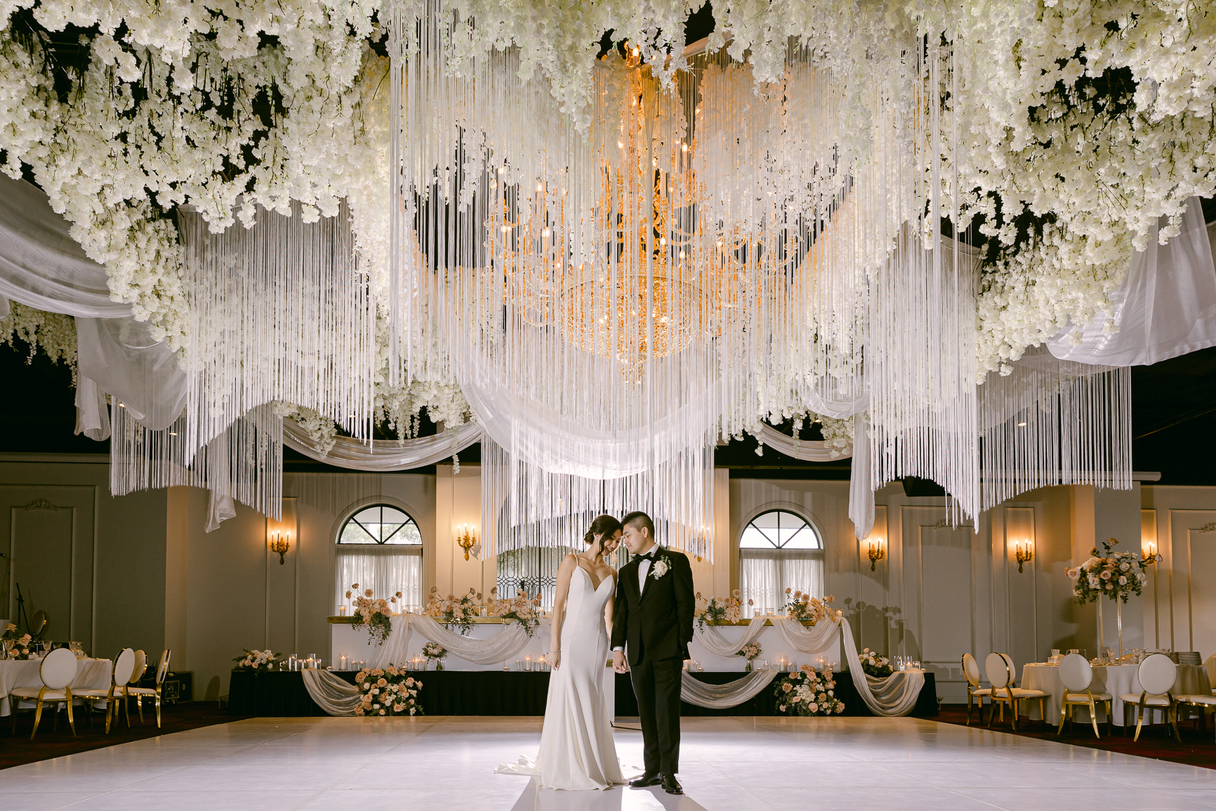 Couple beneath a white floral ceiling installation and chandelier in a ballroom