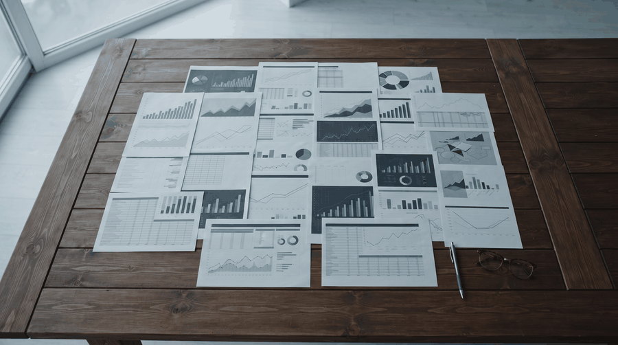 A desk of printed financial charts and reports under a single lamp