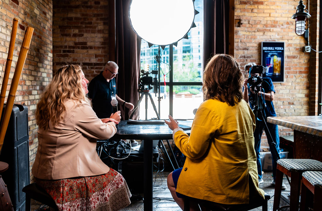 Two guests recorded at a window table for a podcast, with a camera and a soft light beside them