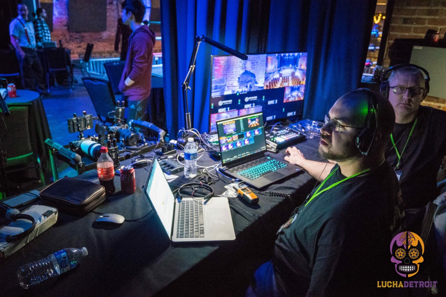 Two operators on headsets at a live switching desk at a Lucha Detroit wrestling show, monitors and mixer lit blue