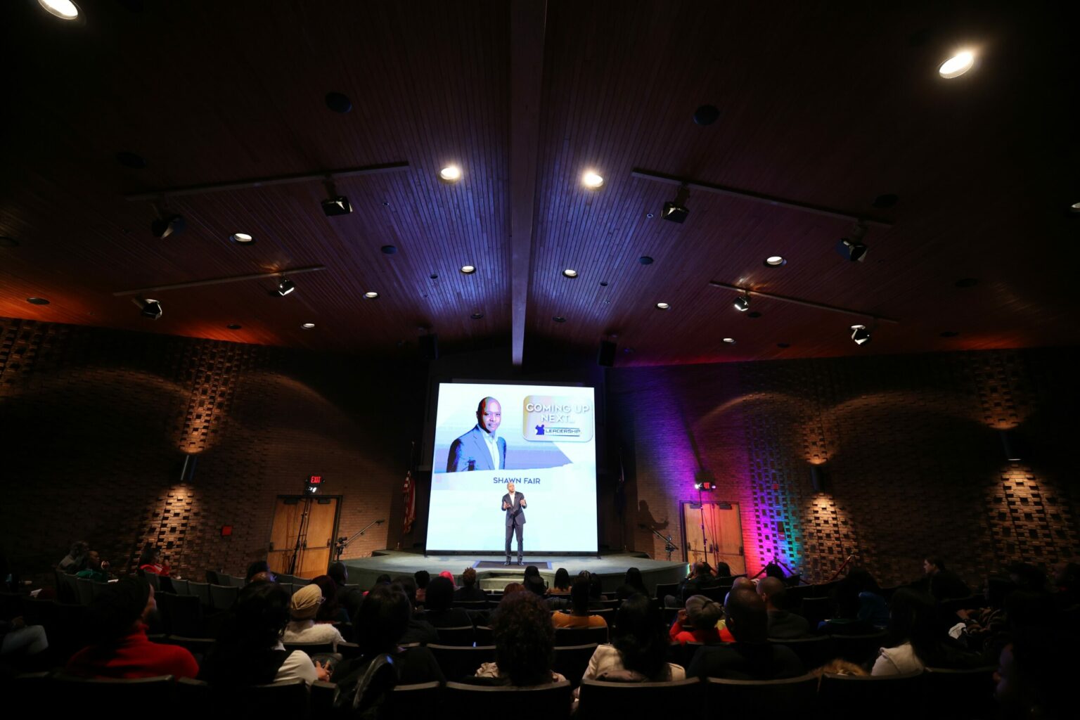 A speaker alone on a dark auditorium stage, a screen lit behind them