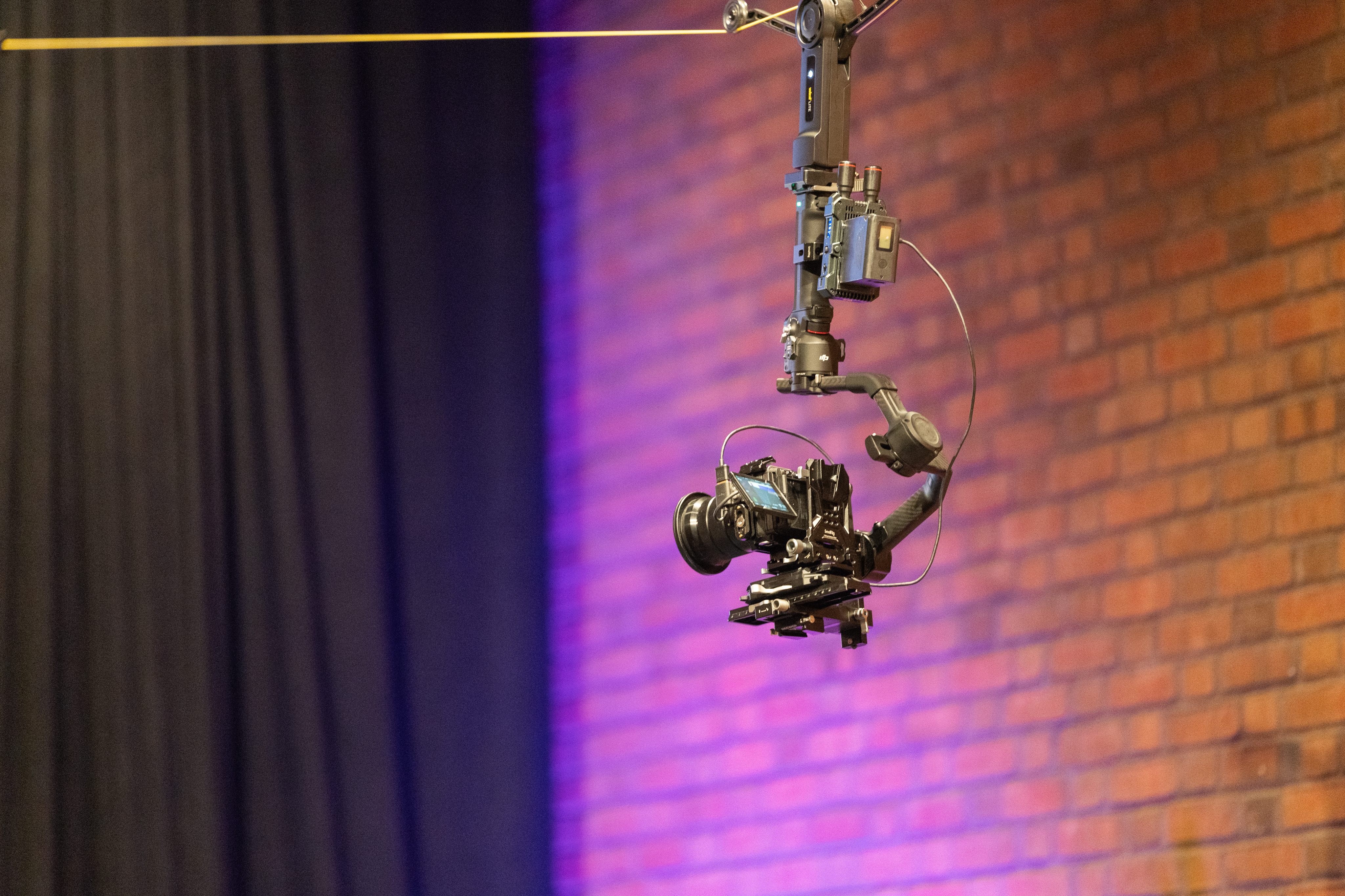 A cinema camera flying on a cable camera rig above a venue floor under purple lighting
