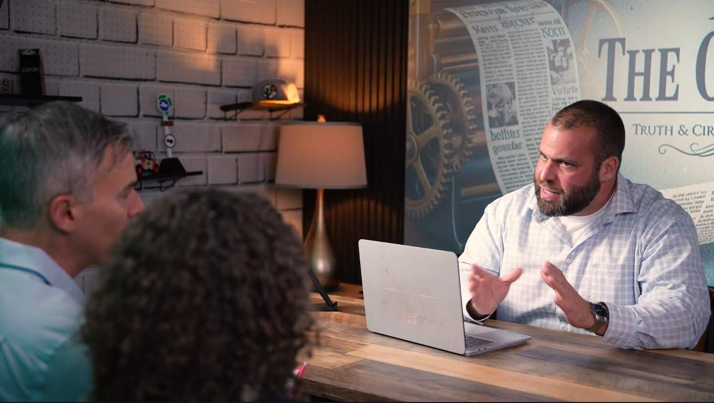 The Gazette, editor's angle: the editor gestures across a laptop at two reporters, the practical brick wall and masthead behind him