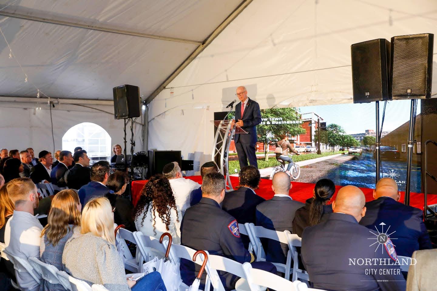 A speaker at a podium addressing seated guests under a tent, with a project rendering on the screen behind