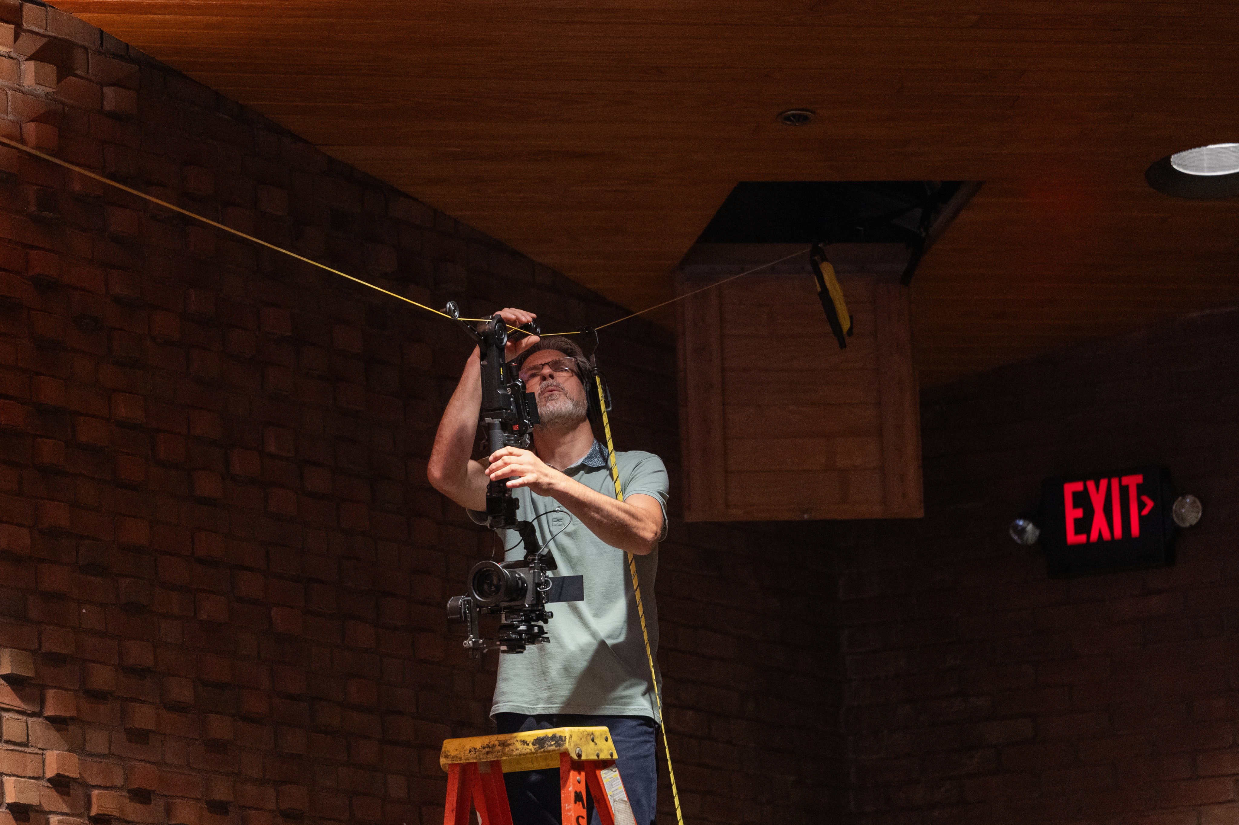 Emil Puste up a ladder rigging a cable camera line across a venue ceiling