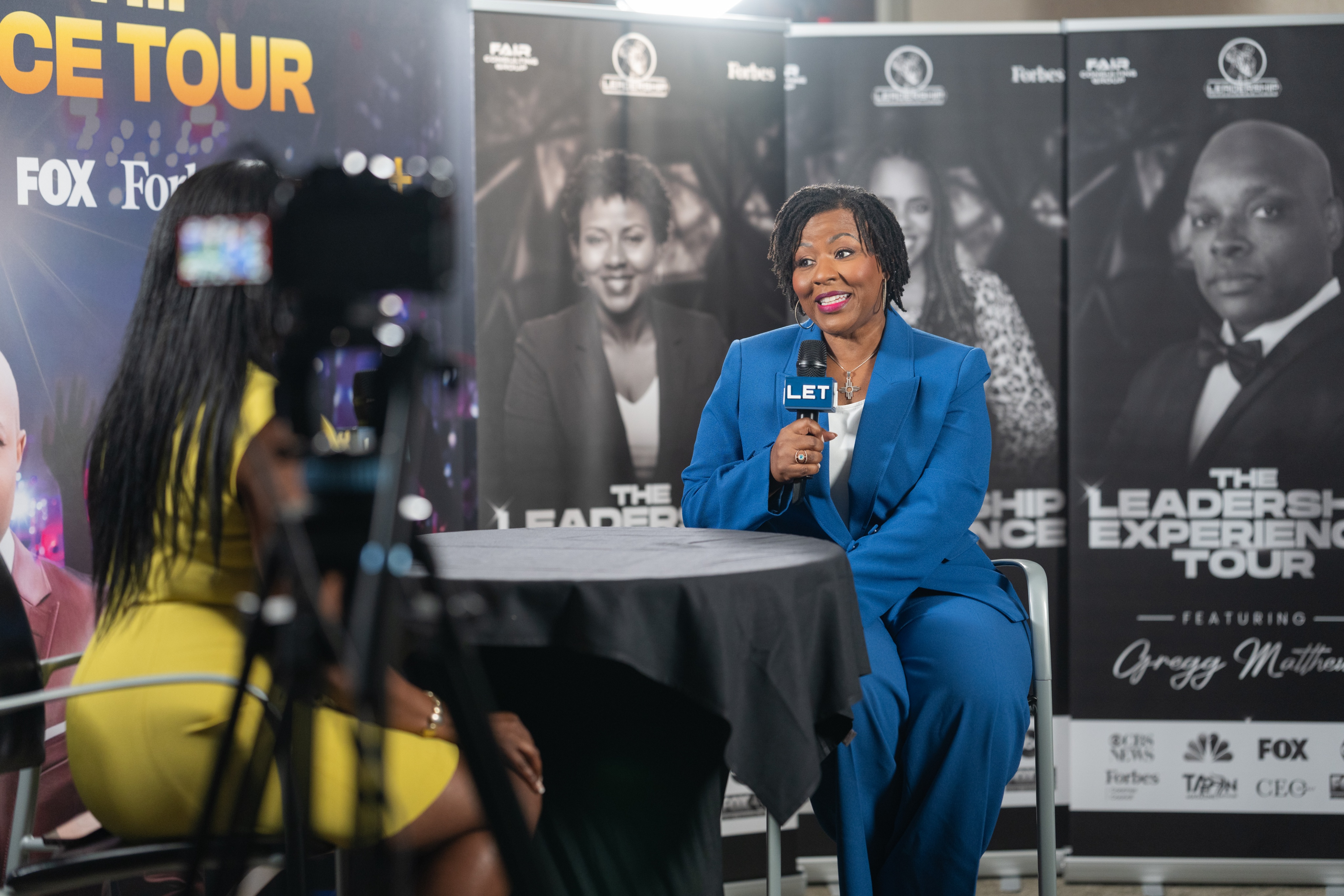 Two guests interviewed on a branded set, seen past a camera