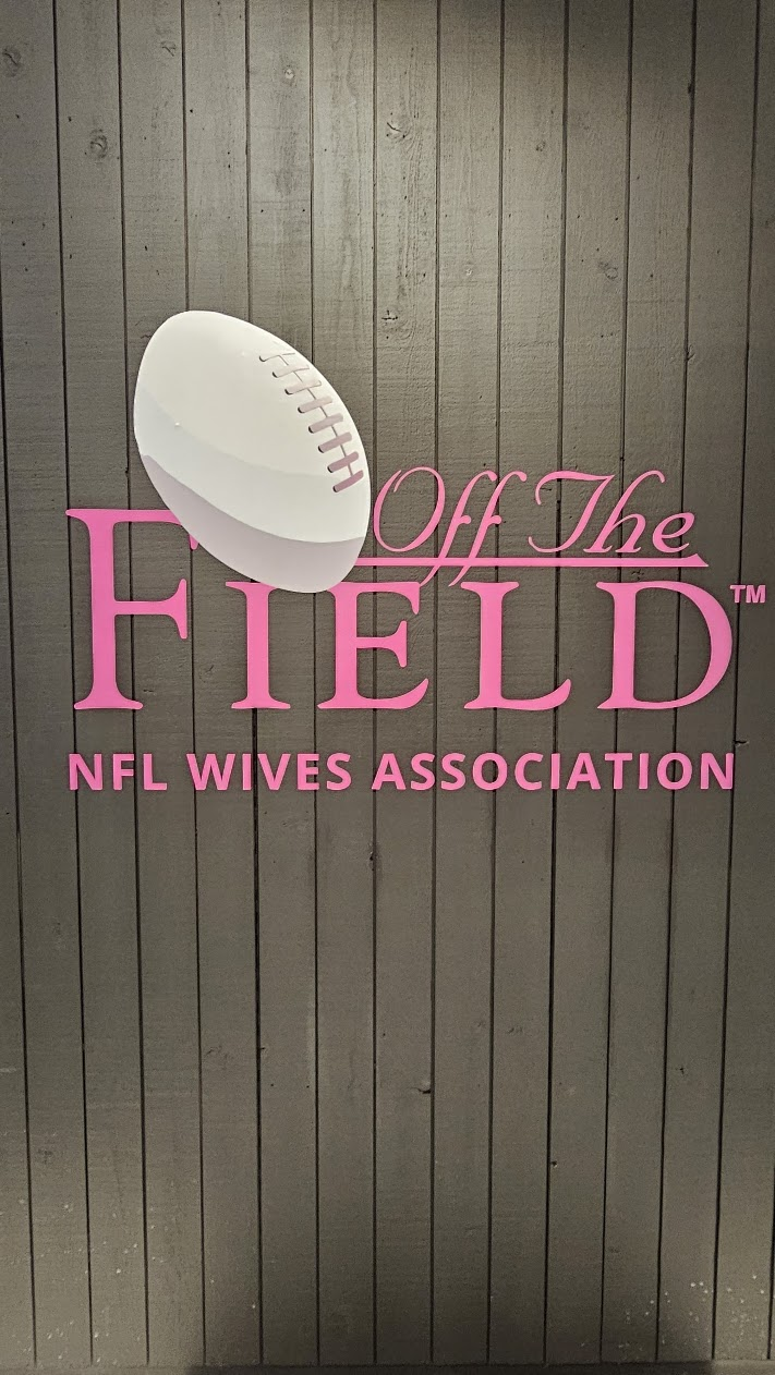 The Off The Field NFL Wives Association sign in pink letters with a white football on a grey wooden wall