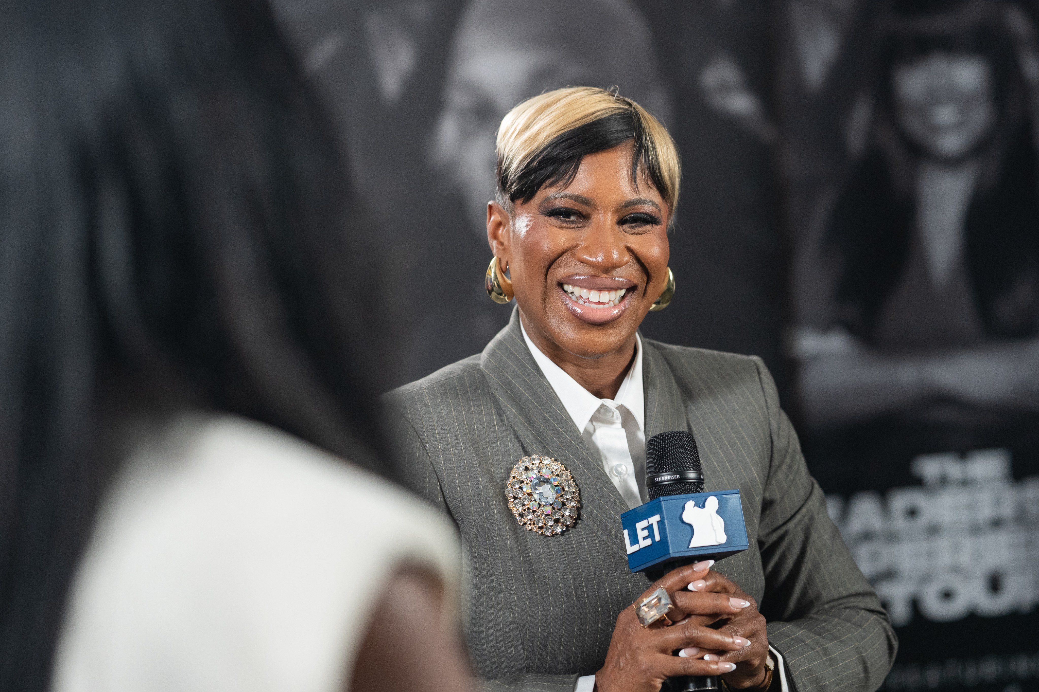 A speaker in a grey suit interviewed on camera holding a branded microphone