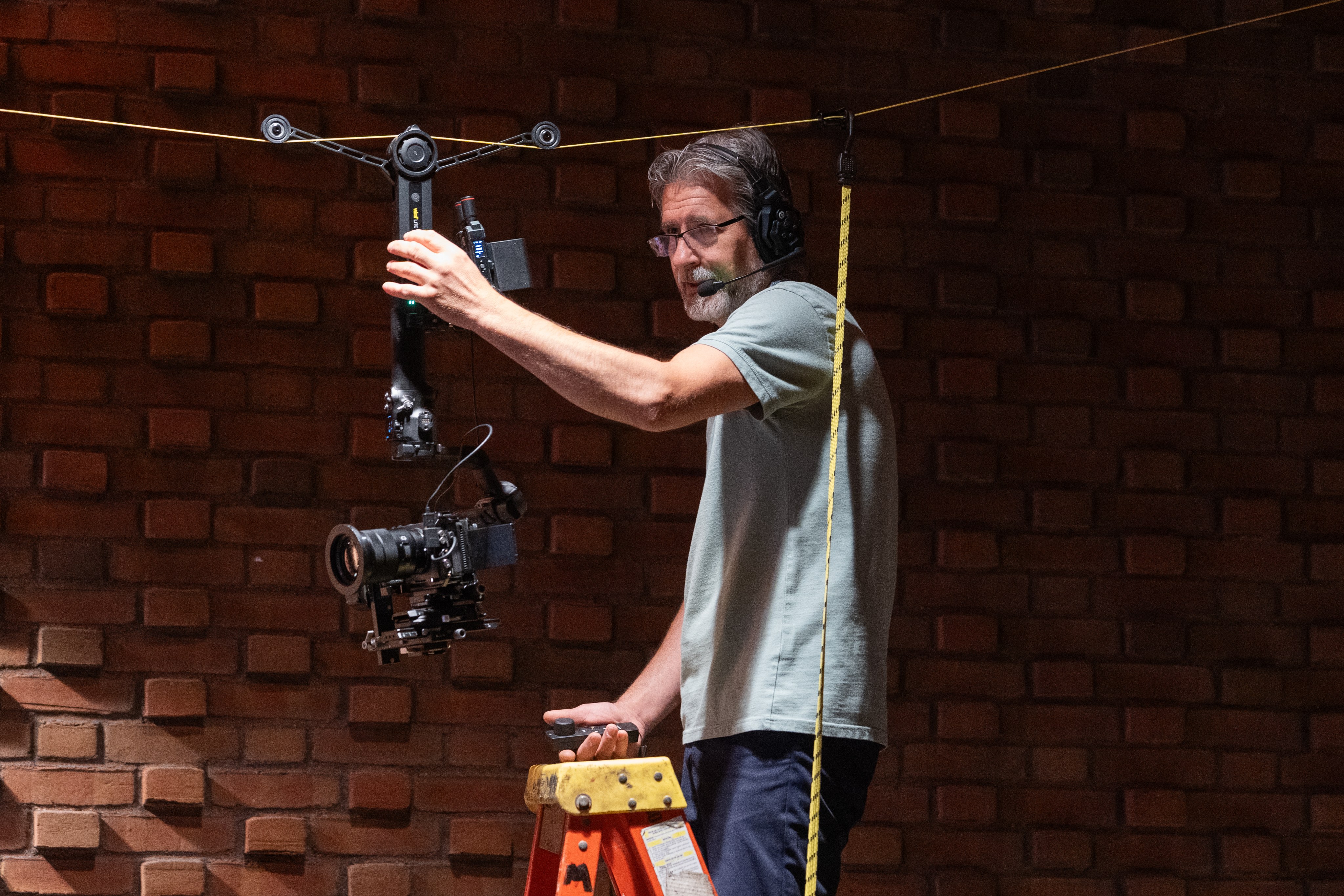 A crew member on a ladder hanging a cable camera line across a venue