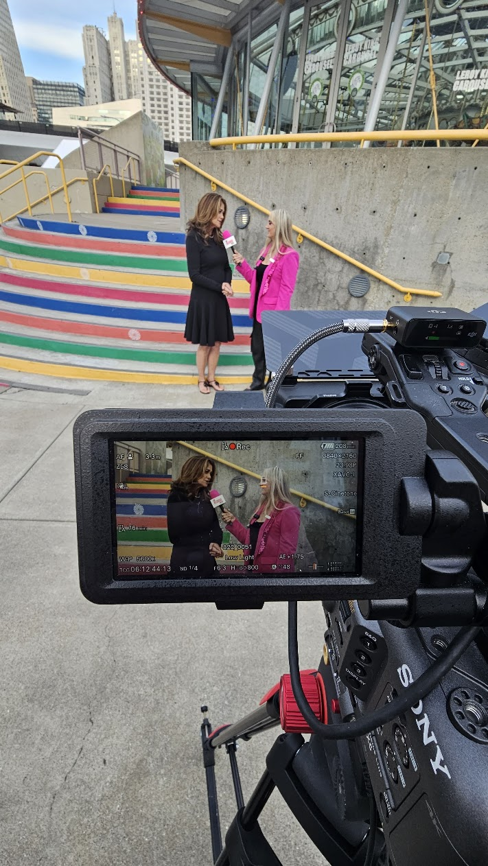 Kathy Ireland being interviewed by a woman in a pink blazer, framed on a camera's monitor beside multicoloured outdoor steps