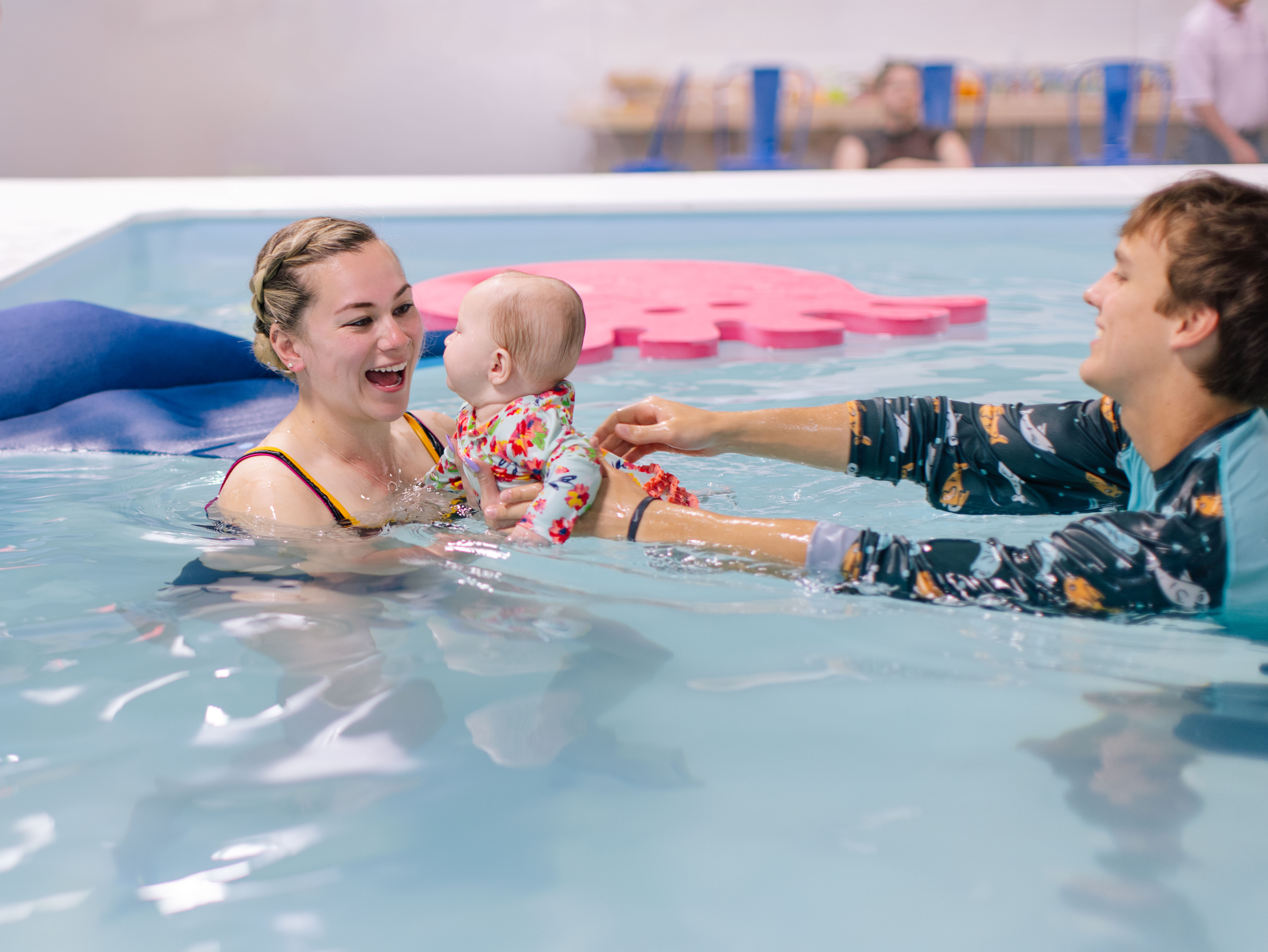 Blonde instructor laughing as a colleague hands her a baby in the pool