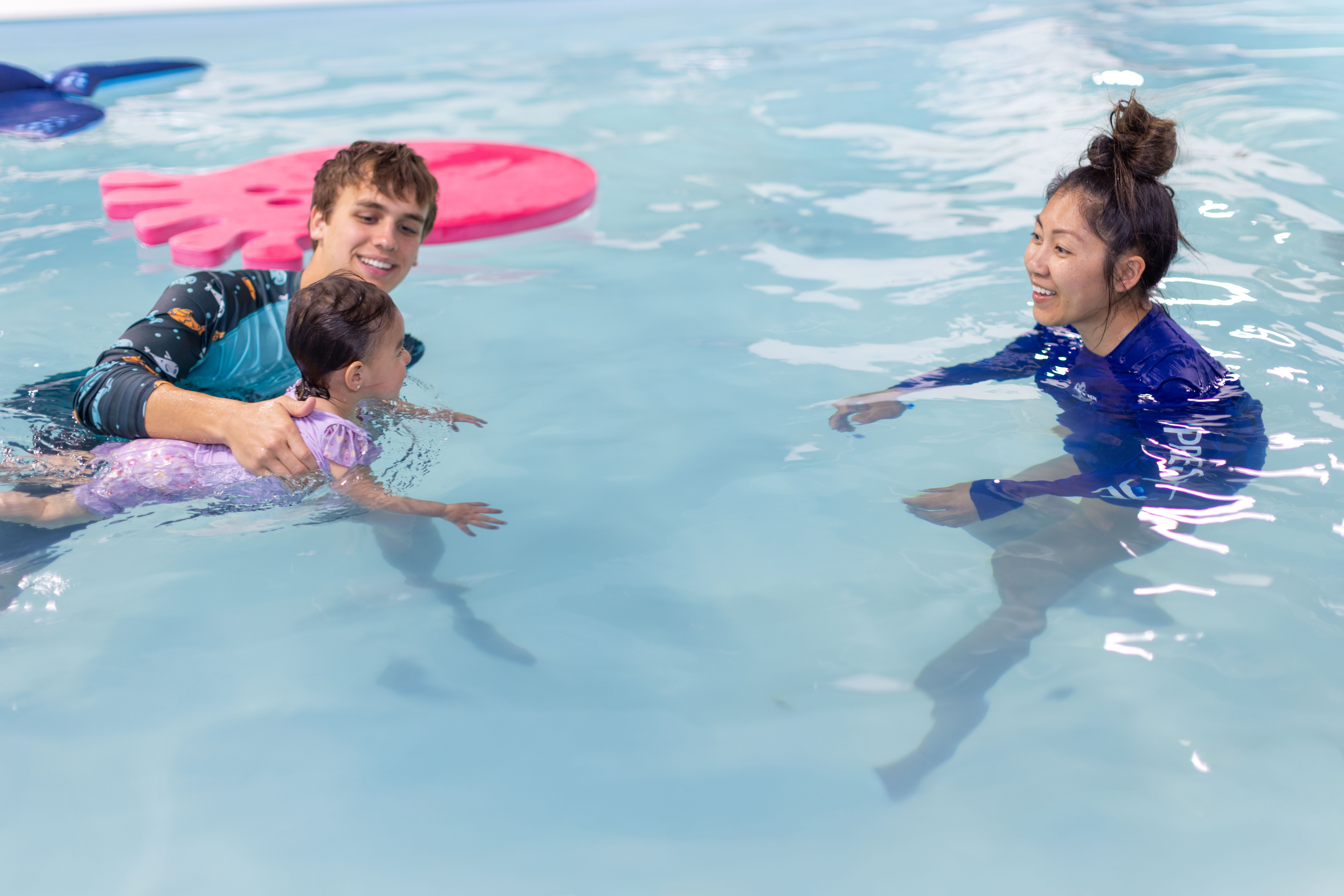 Two instructors work together: one holds a baby in a float position while the other extends a hand