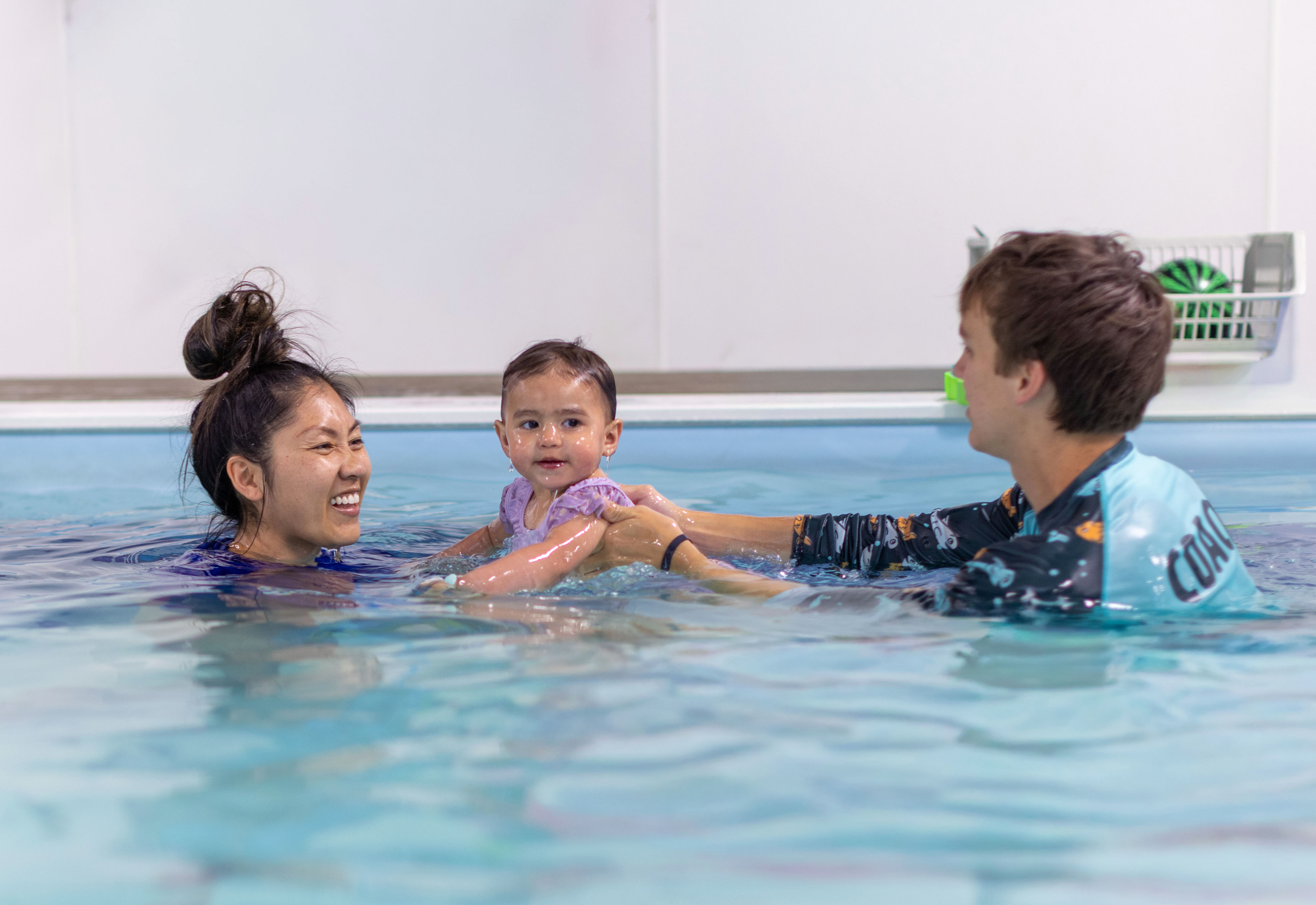 Two instructors laughing together as they support a dark-haired swimmer