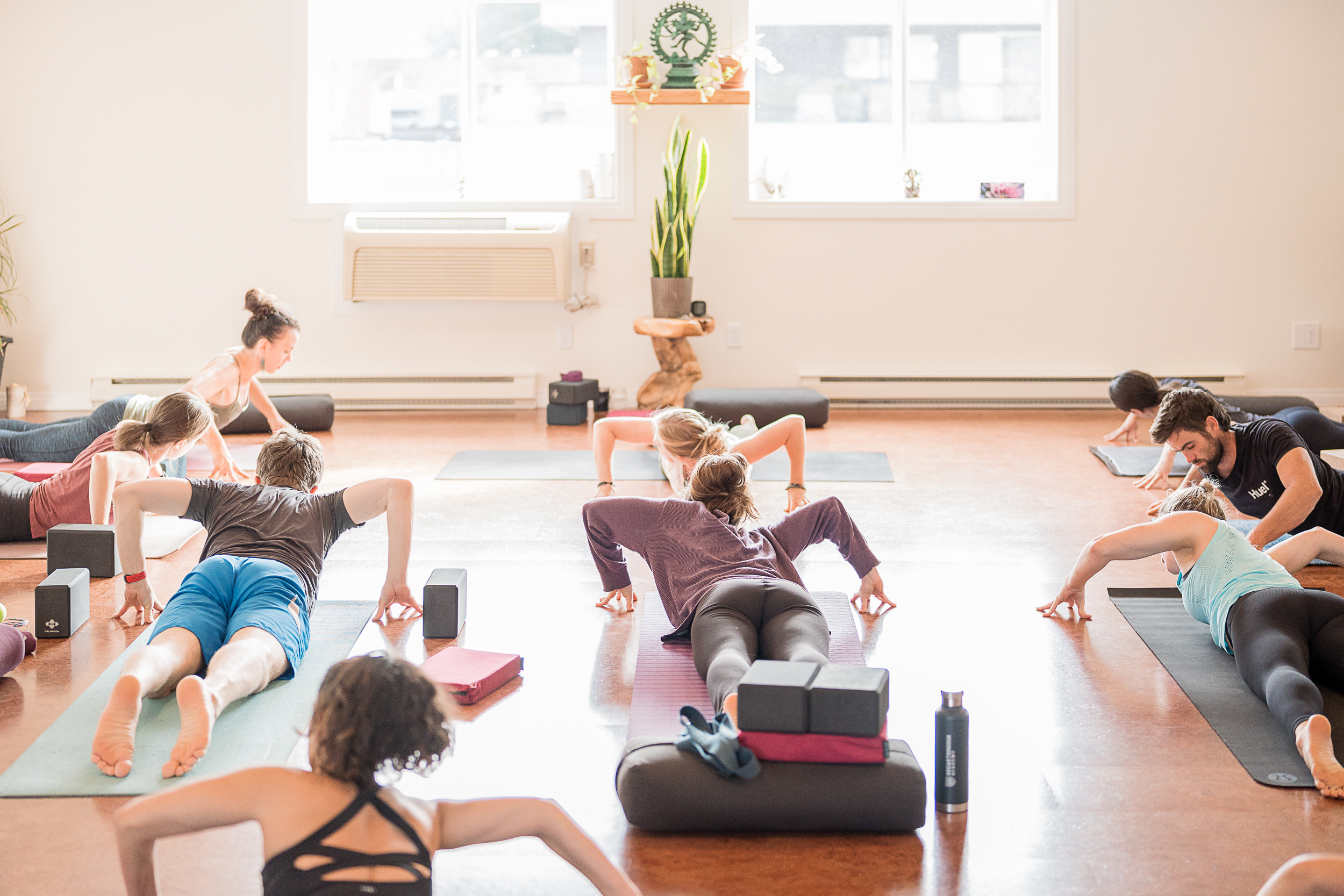 Yoga practitioners in a bright, airy Shala Yoga studio in Squamish engage in a relaxing floor stretch with support blocks, under the guidance of a teacher, promoting wellness and community spirit.