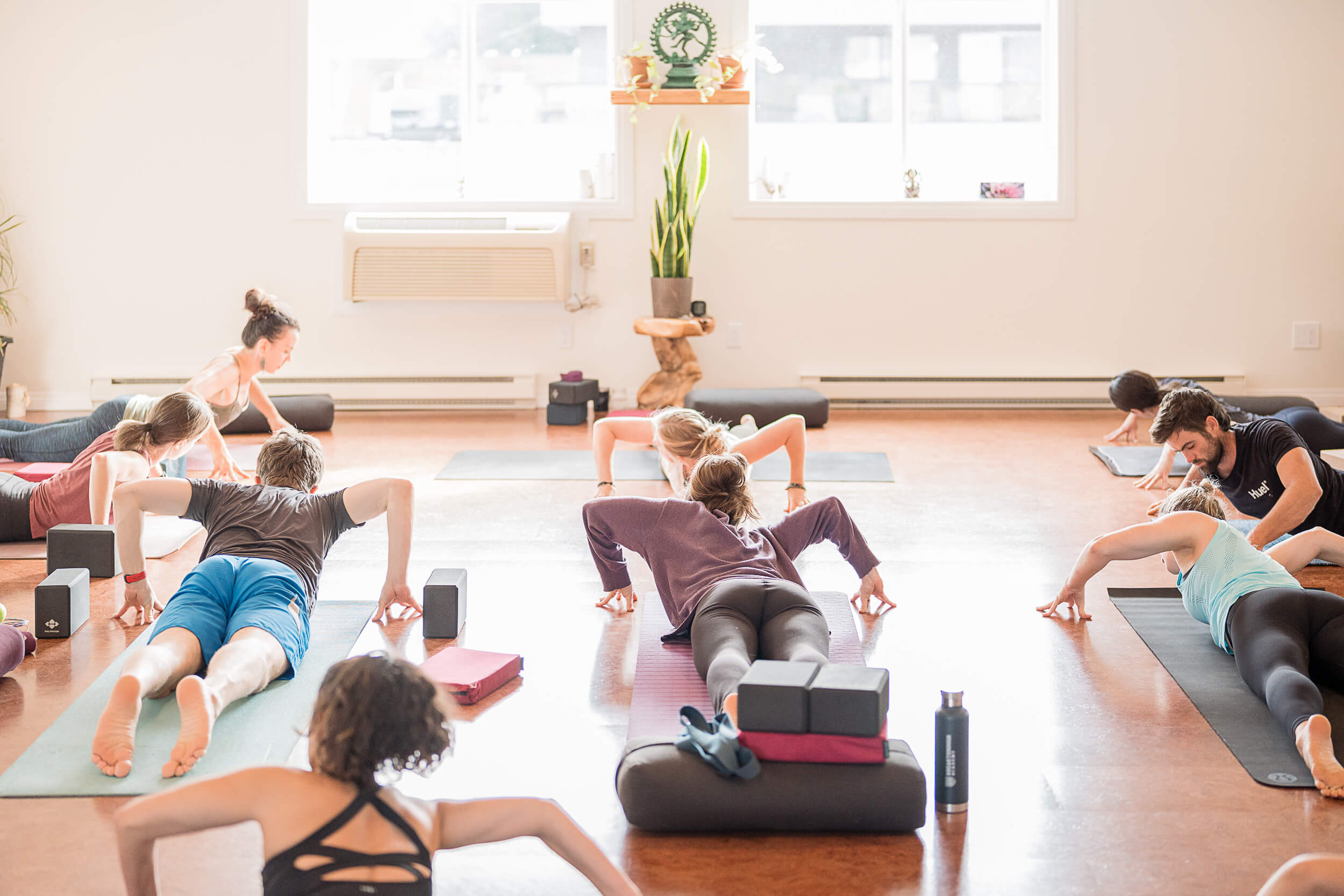 Diverse group of individuals practicing a yoga stretch in a sunlit studio, with yoga mats, blocks, and water bottles, embodying the essence of wellness and community in Squamish yoga studio.