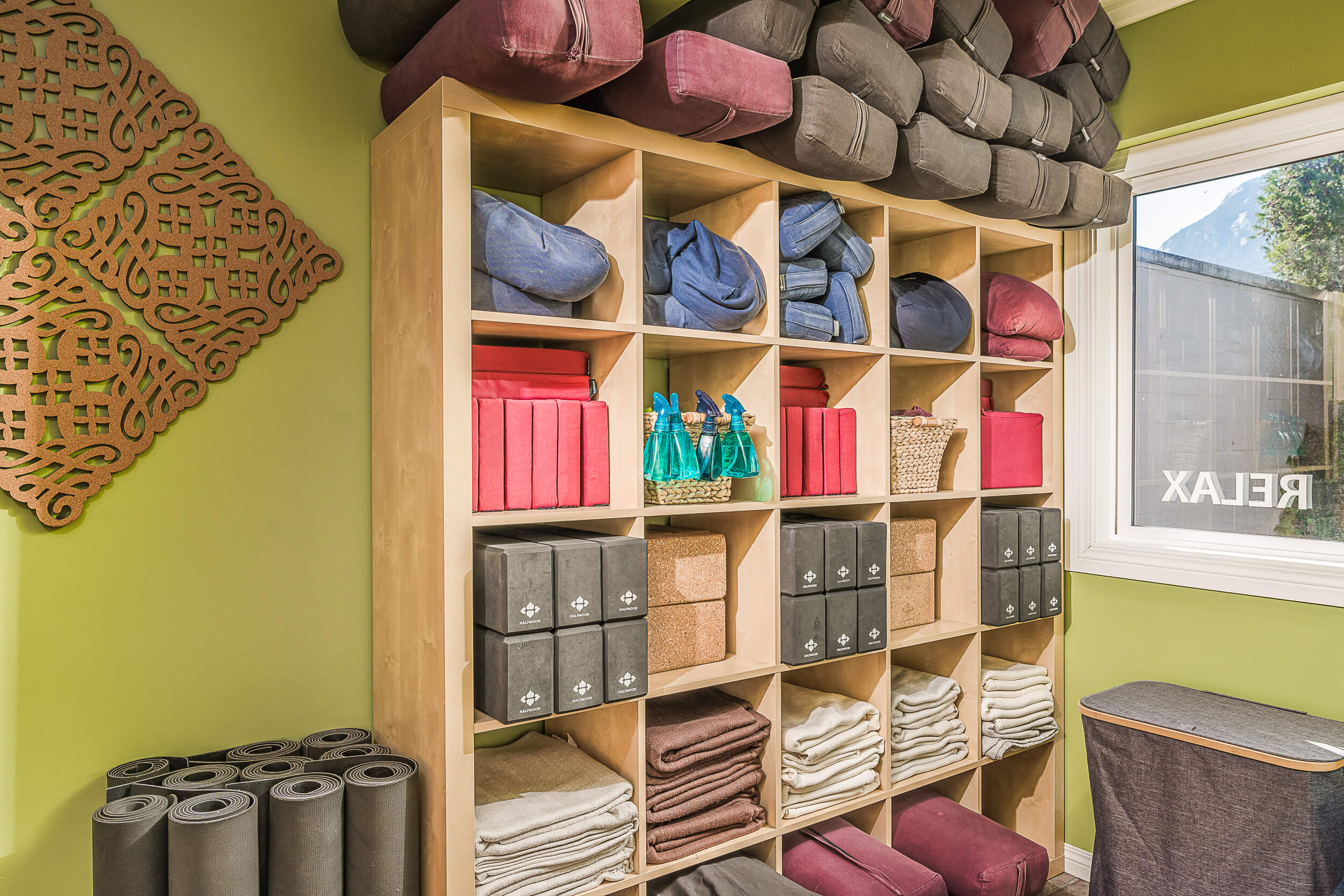 Well-organized yoga props on wooden shelves at Shala Yoga studio in Squamish, featuring an assortment of colorful bolsters, neatly folded blankets, and cork blocks, complemented by a decorative wooden mandala on a lime green wall, enhancing the serene and welcoming atmosphere for practitioners.