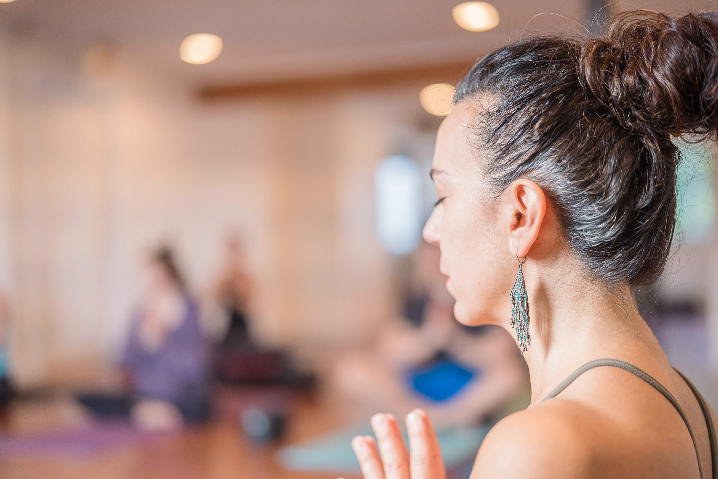 Close-up of a yoga instructor's profile with her hand in Anjali Mudra, eyes closed in concentration, as other participants practice in the softly-focused background at Shala Yoga studio in Squamish.