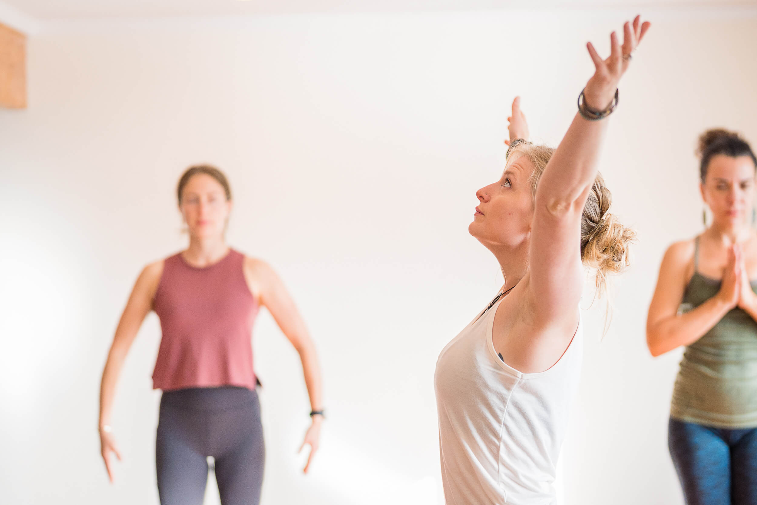 Focused yoga students at Shala Yoga studio in Squamish performing a standing stretch with one arm raised, embodying balance and flexibility, while another participant practices Anjali Mudra in the background, showcasing the variety of poses in a yoga sequence.