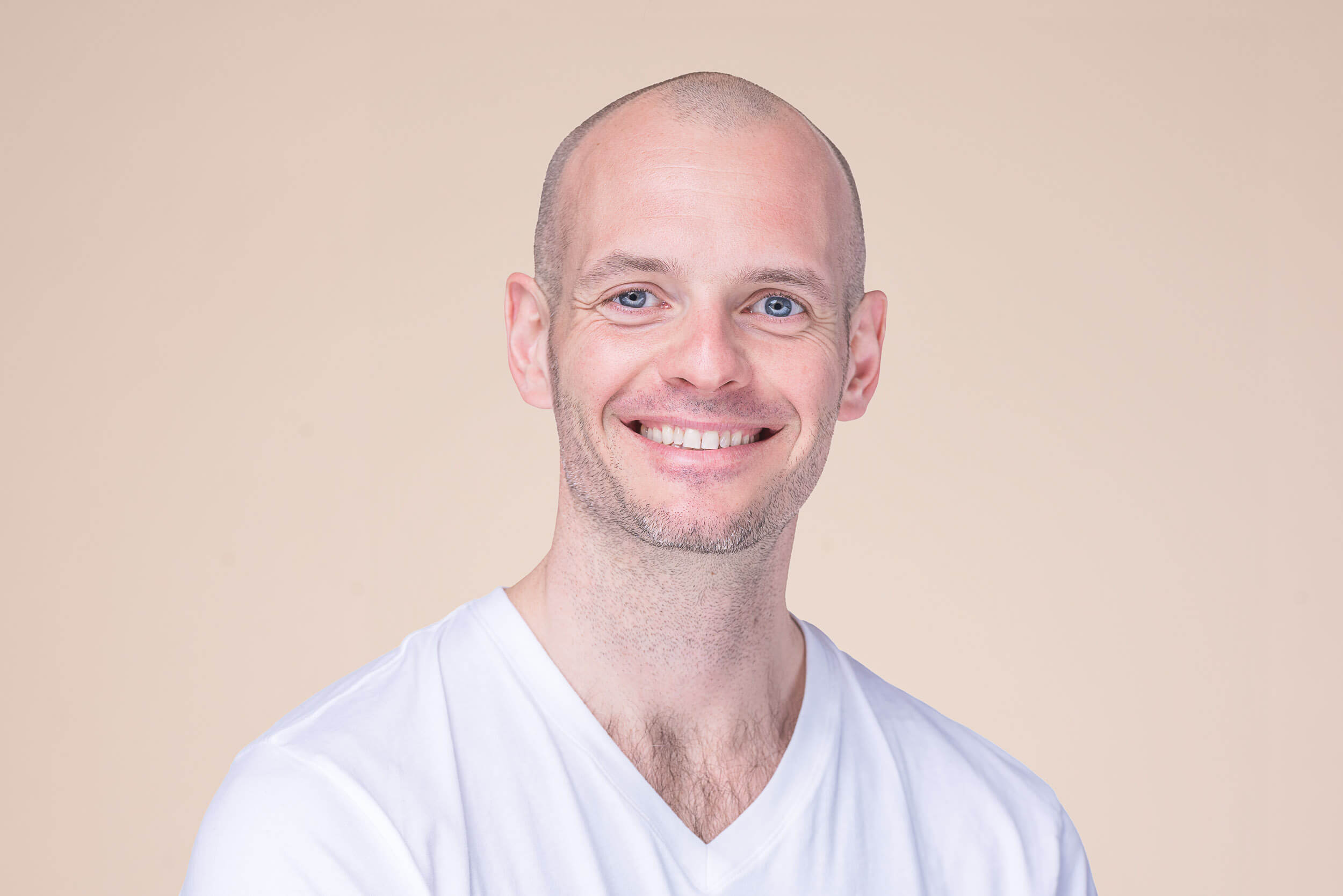 Friendly male yoga instructor with a bald head and bright smile, wearing a white V-neck t-shirt, representing the approachable and professional staff at Shala Yoga studio in Squamish.