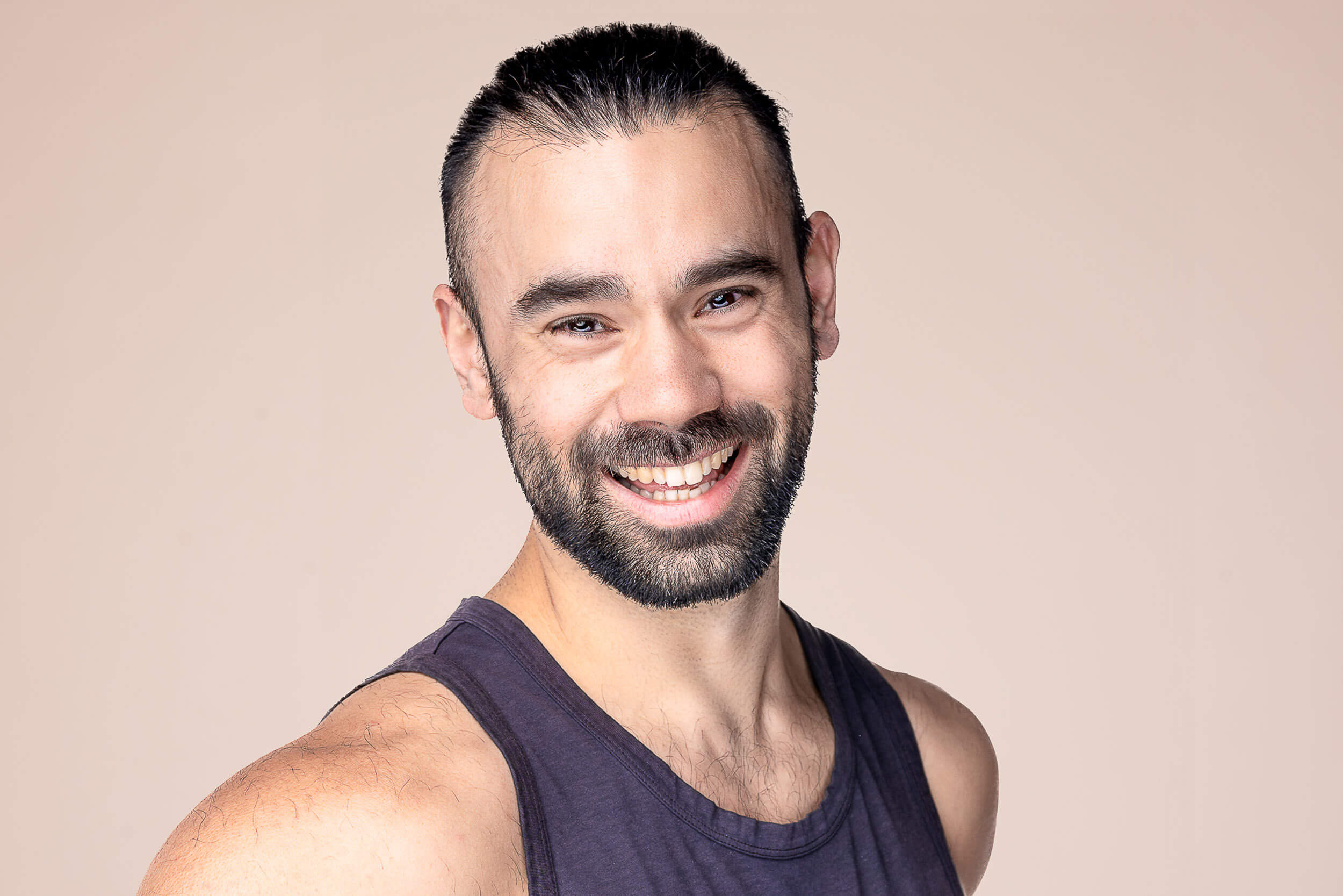 Energetic and friendly male yoga instructor with a beard, wearing a sleeveless dark tank top and a warm smile, epitomizing the inviting and positive energy at Shala Yoga studio in Squamish.