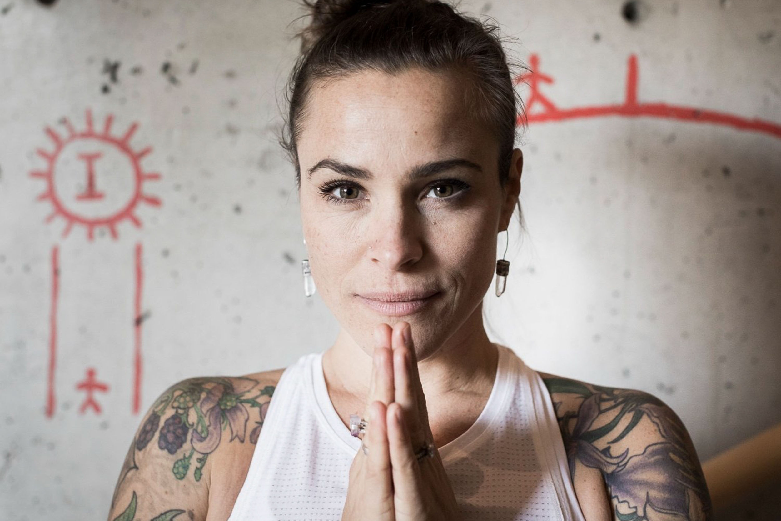 A focused yoga instructor at Shala Yoga studio in Squamish assumes the Anjali Mudra pose, her tattoos creating a unique contrast with the spiritual symbol on the wall behind her, embodying the studio's embrace of individuality and inner peace.