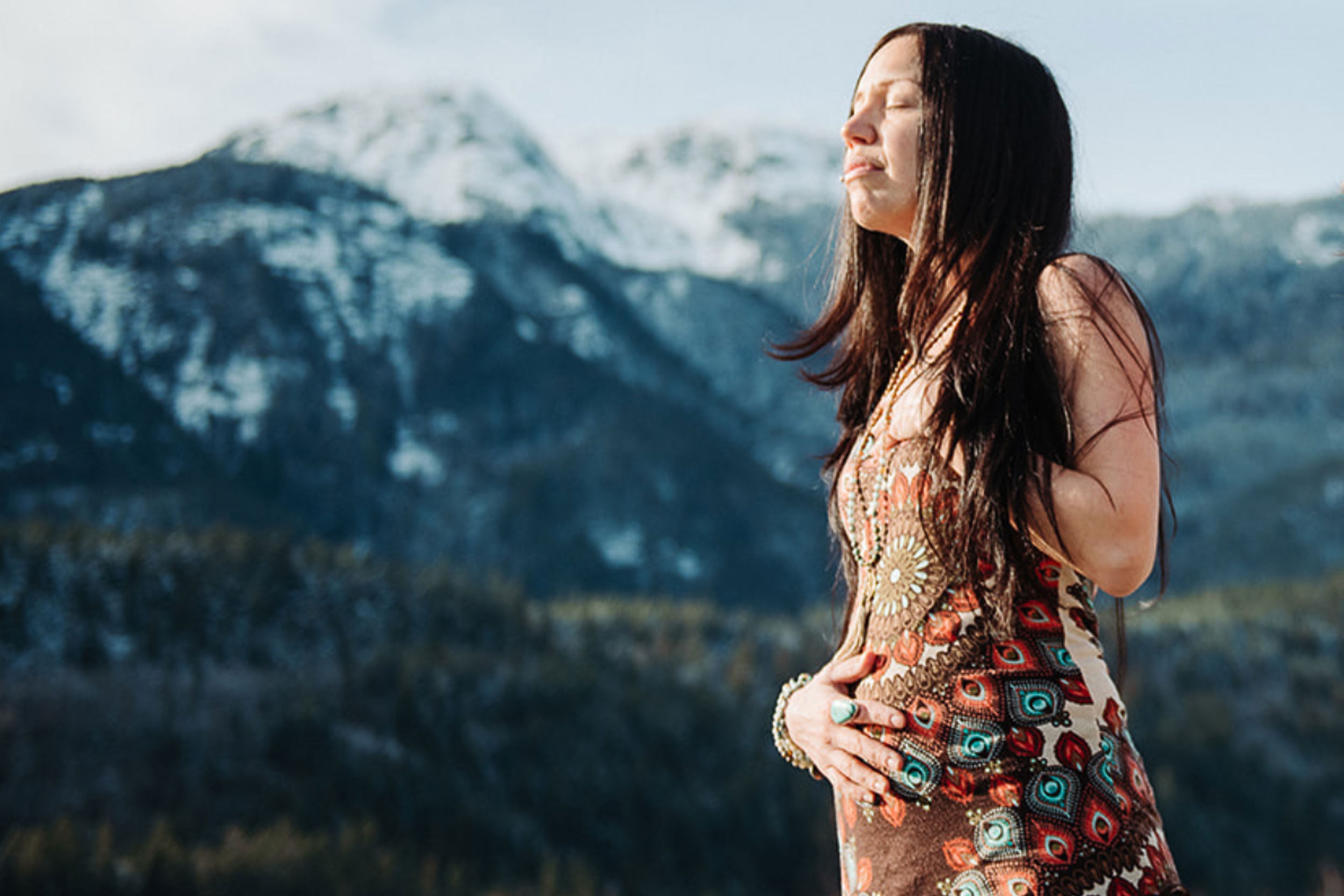 Woman in a contemplative pose wearing a patterned dress, standing against the backdrop of the majestic snow-capped mountains near Shala Yoga studio in Squamish, reflecting the studio's commitment to harmony with the natural world.