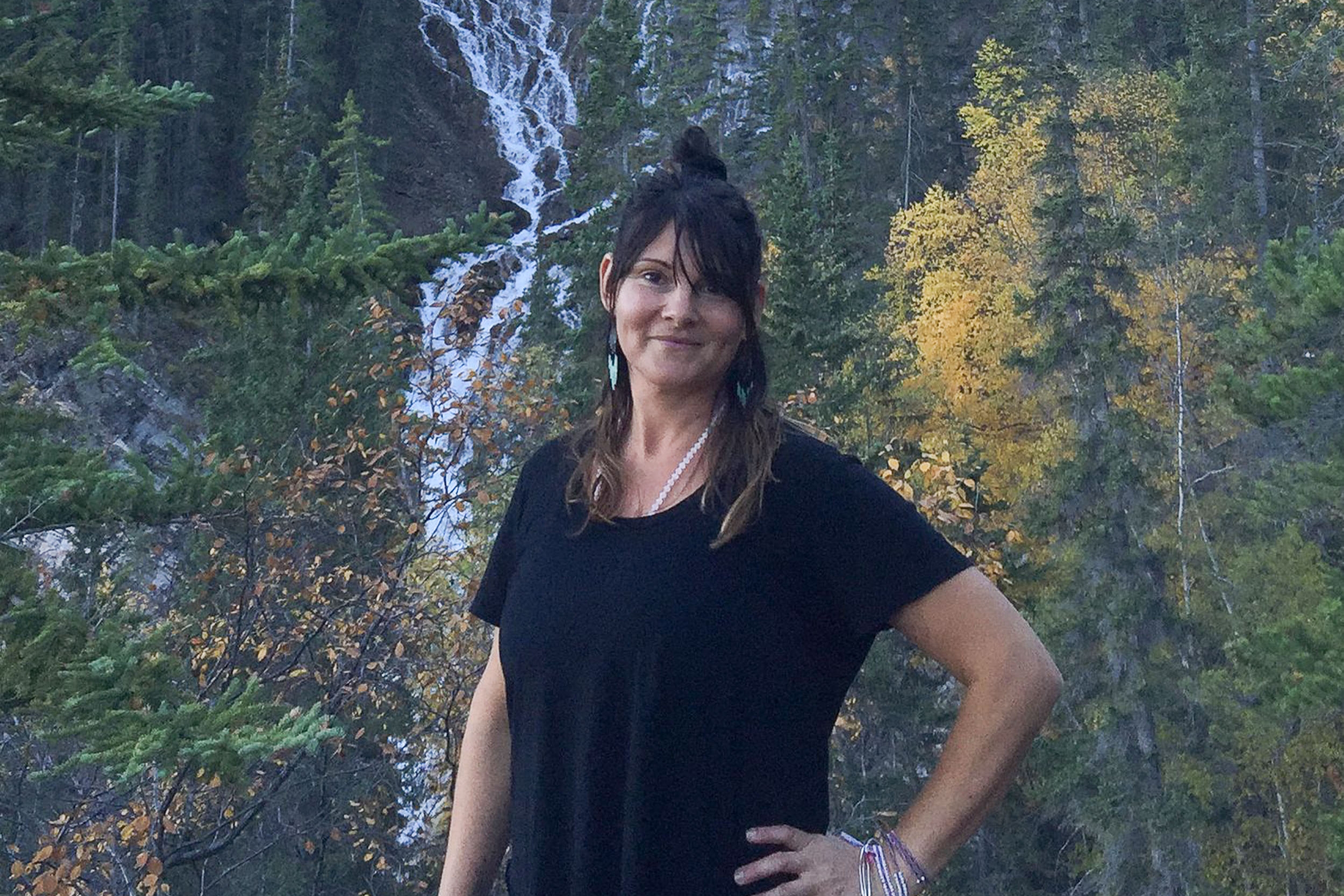 Confident yoga instructor in a casual black outfit standing in front of a waterfall surrounded by evergreens and autumn foliage, embodying the connection with nature that Shala Yoga studio in Squamish encourages.
