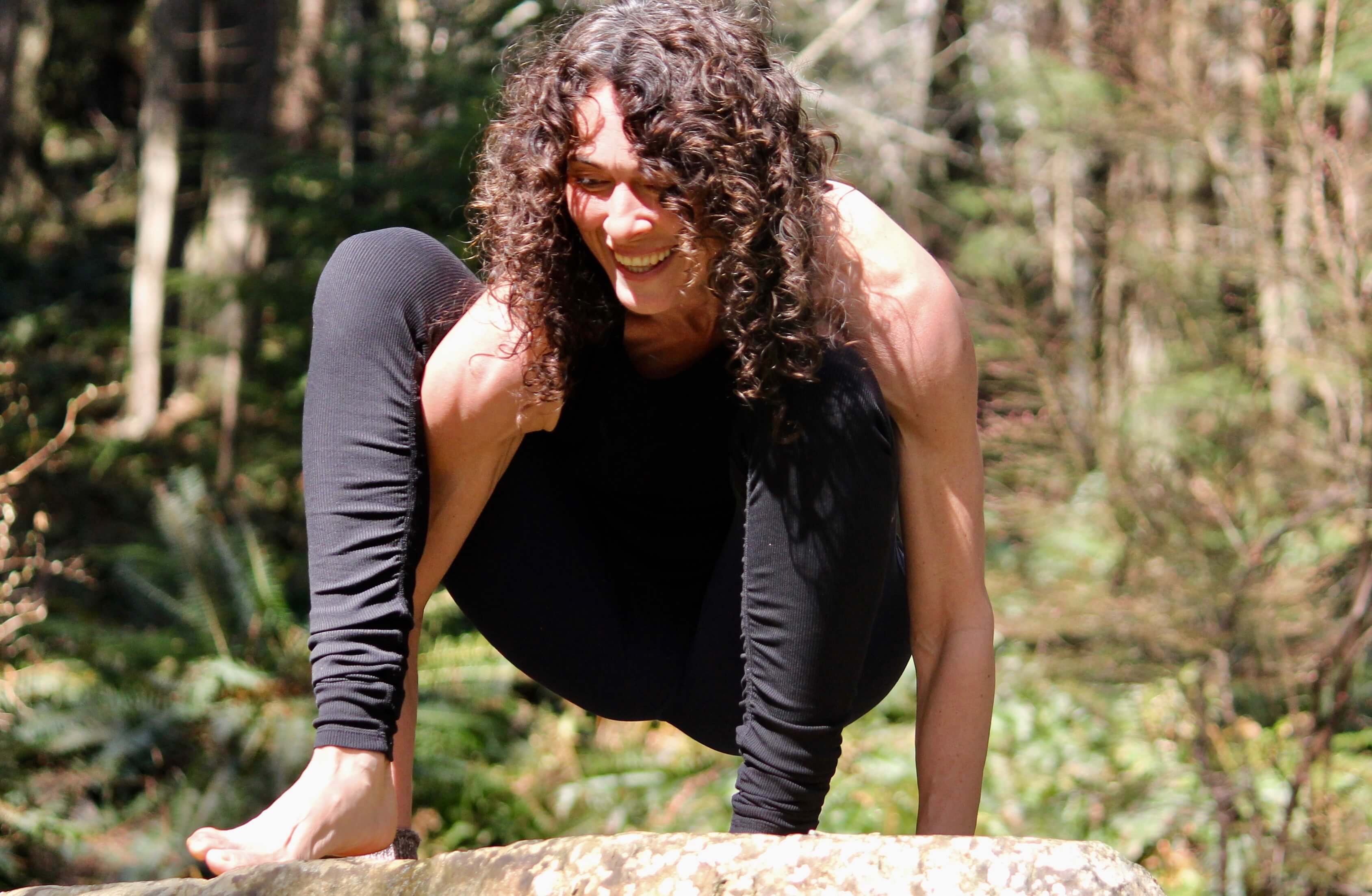 Yoga practitioner in a black outfit performing an arm-balanced pose on a rock outdoors, her laughter blending with the natural surroundings, showcasing the joy of yoga practice with Shala Yoga studio in Squamish.