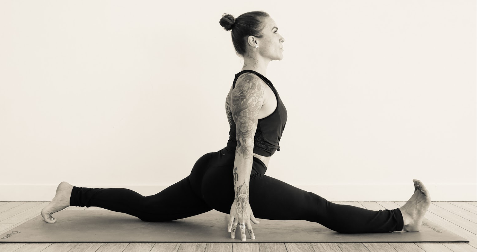 Monochrome image of a dedicated yoga instructor demonstrating an advanced split pose at Shala Yoga studio in Squamish, her tattoos adding to the artistic expression of strength and flexibility