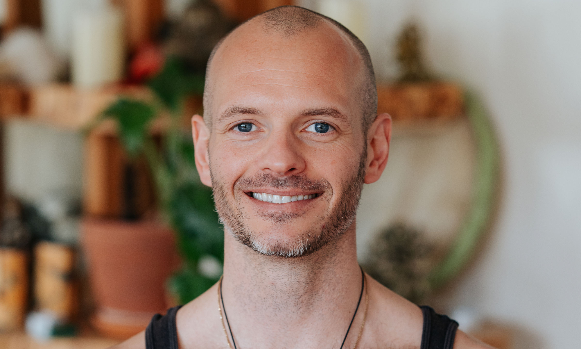 Friendly male yoga instructor with a bald head and bright smile, wearing a white V-neck t-shirt, representing the approachable and professional staff at Shala Yoga studio in Squamish.