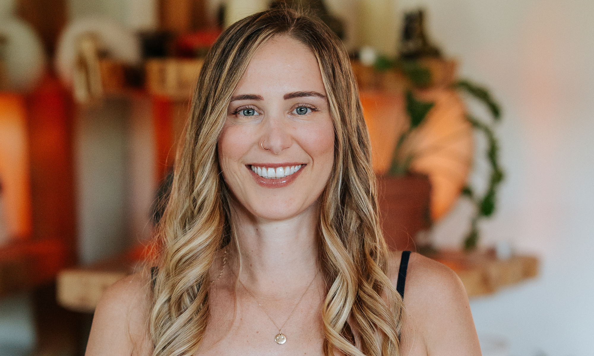 Warm and welcoming yoga instructor with a gentle smile, wearing a simple black top, representing the accessible and friendly environment of Shala Yoga studio in Squamish.