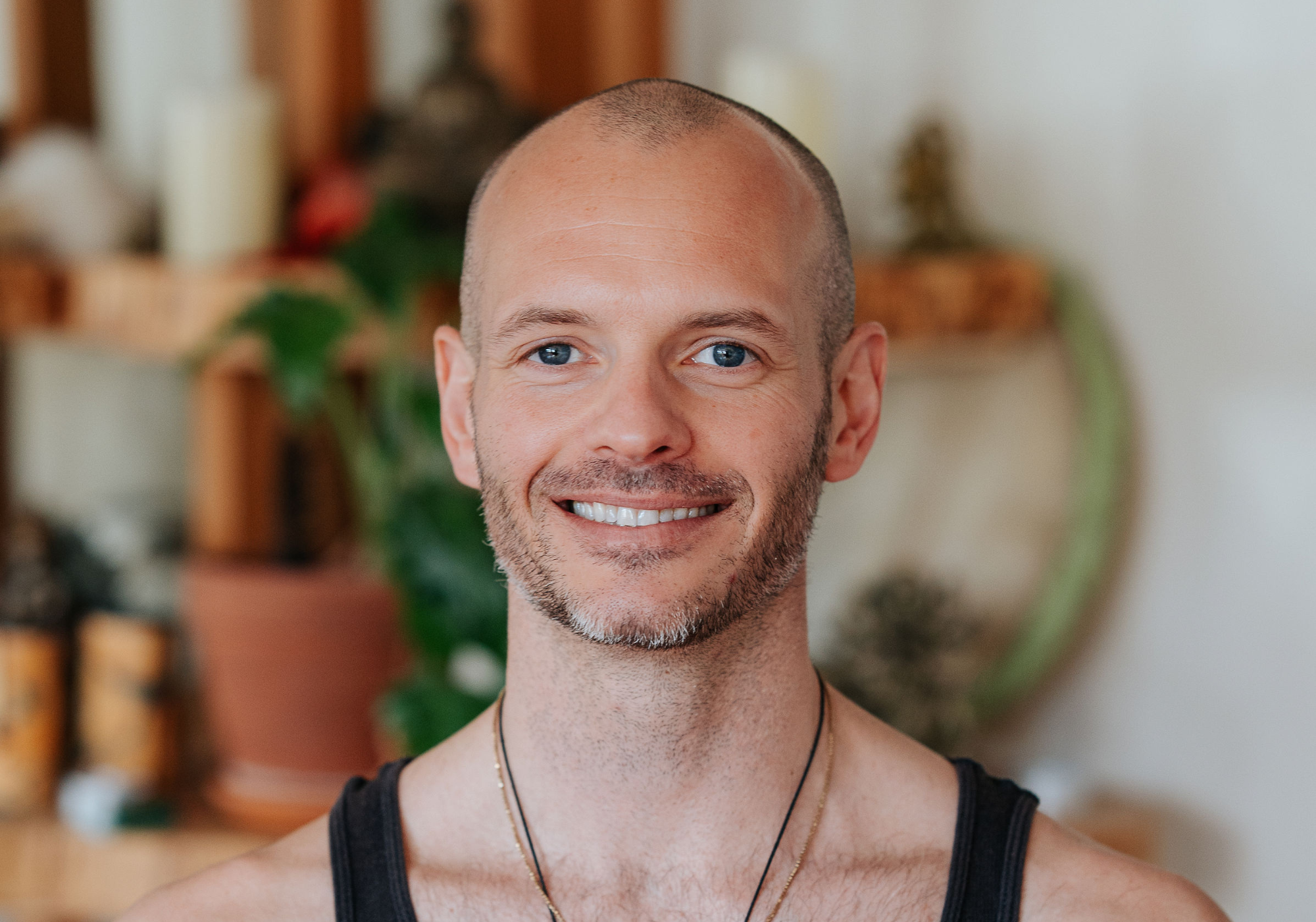 Friendly male yoga instructor with a bald head and bright smile, wearing a white V-neck t-shirt, representing the approachable and professional staff at Shala Yoga studio in Squamish.