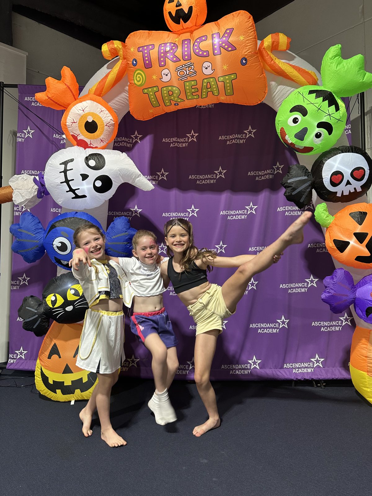Three kids in costume striking fun poses under a giant inflatable Trick or Treat arch at the studio