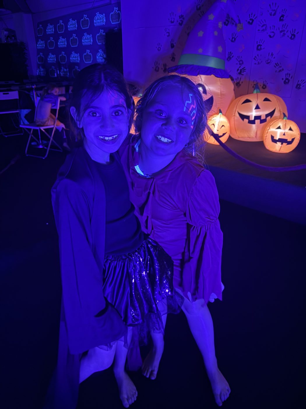 Two smiling girls with glowing face paint under UV lights beside light-up pumpkins