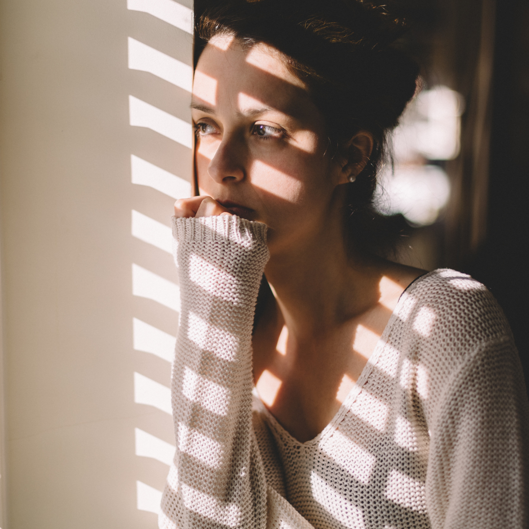 Woman leaning against a wall by a window with light and shadow from blinds across her face, looking thoughtful Woman leaning against a wall by a window with light and shadow from blinds across her face, looking thoughtful