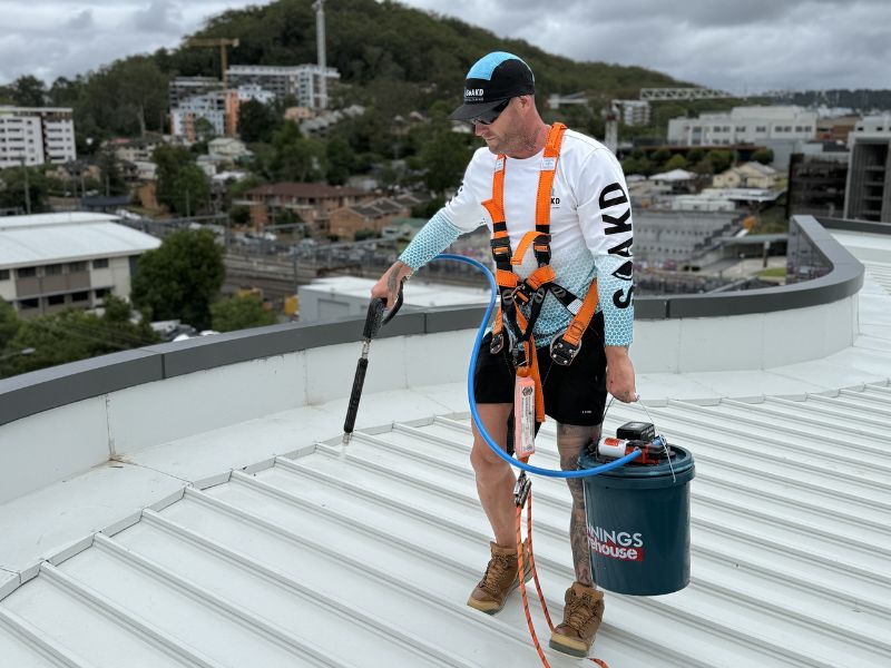 SOAKD operator in full harness and fall-protection gear applying softwash treatment on a commercial metal roof