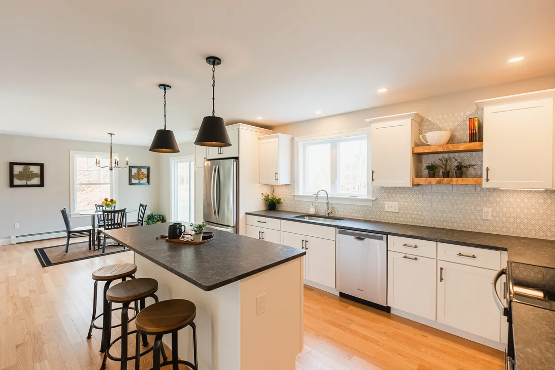 Bright white kitchen with a dark stone island, black pendant lights, wood bar stools and an open dining area beyond
