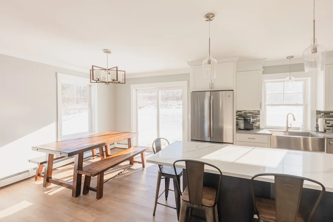 Bright open kitchen with a white quartz island, metal bar stools, a stainless fridge and farmhouse sink, and a wooden trestle dining table with benches beside a sliding glass door