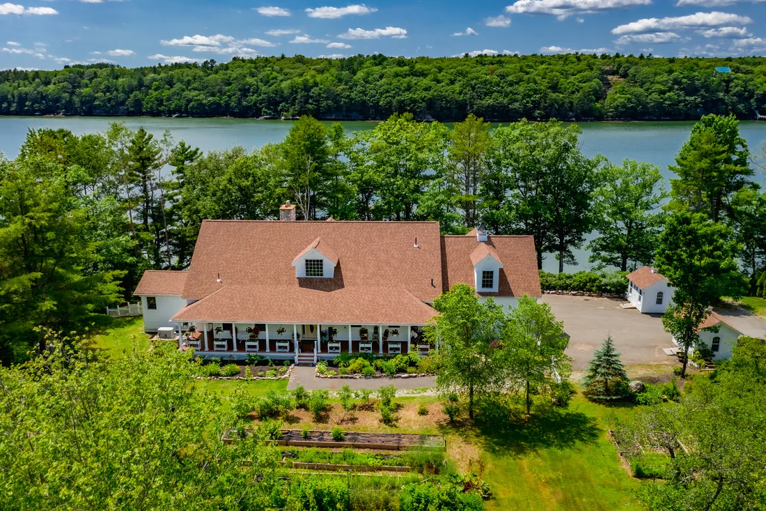 Aerial view of a large white home with a broad roof, dormers and a full front porch, set among tall pines with a wide river and a wooded far shore behind it