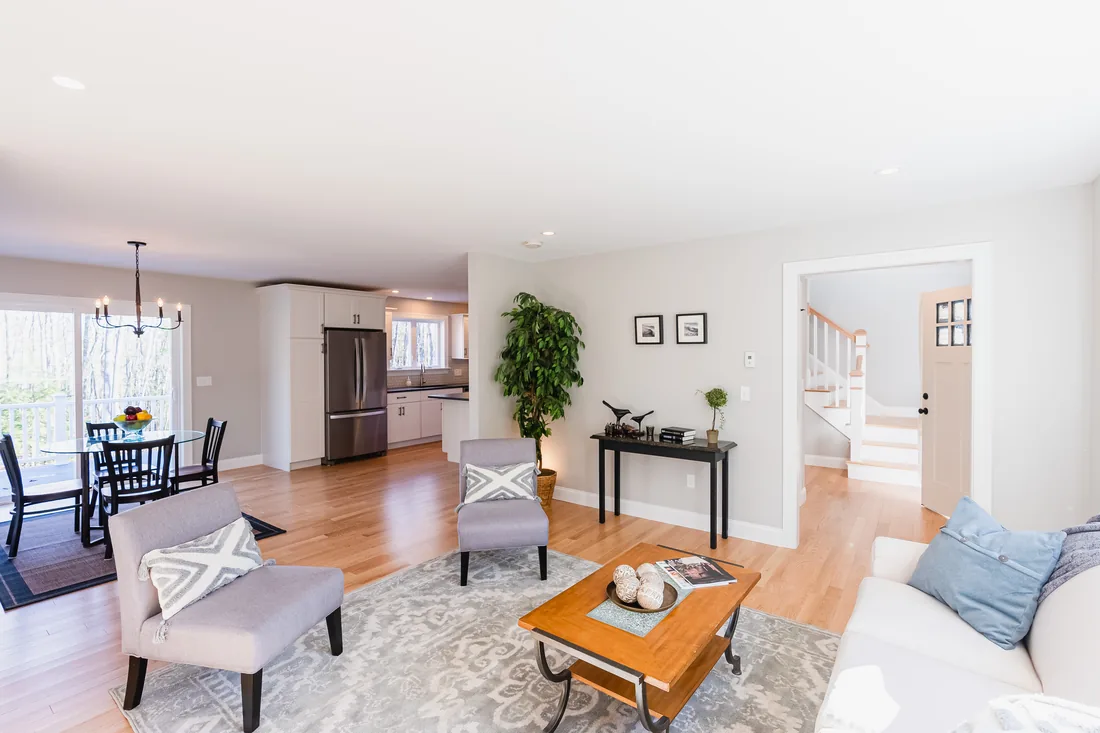 Staged living room with gray armchairs, a white sofa and a wood coffee table, opening to a dining area, a white kitchen and a staircase
