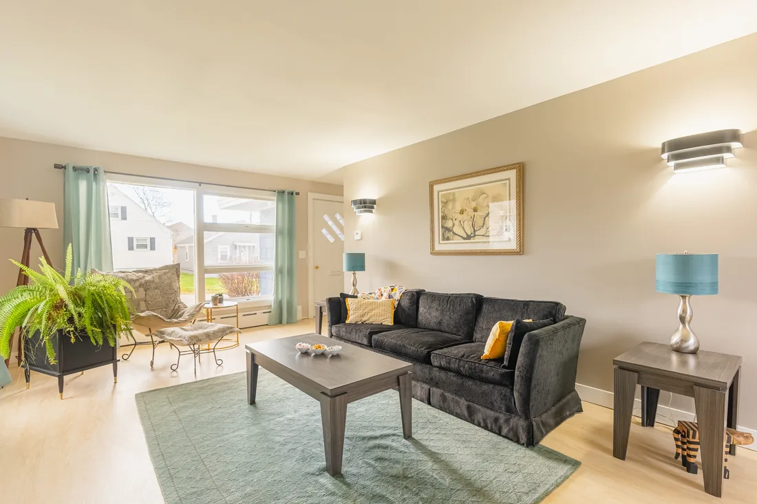Staged living room with a charcoal velvet sofa, a gray coffee table, teal lamps and a wide front window