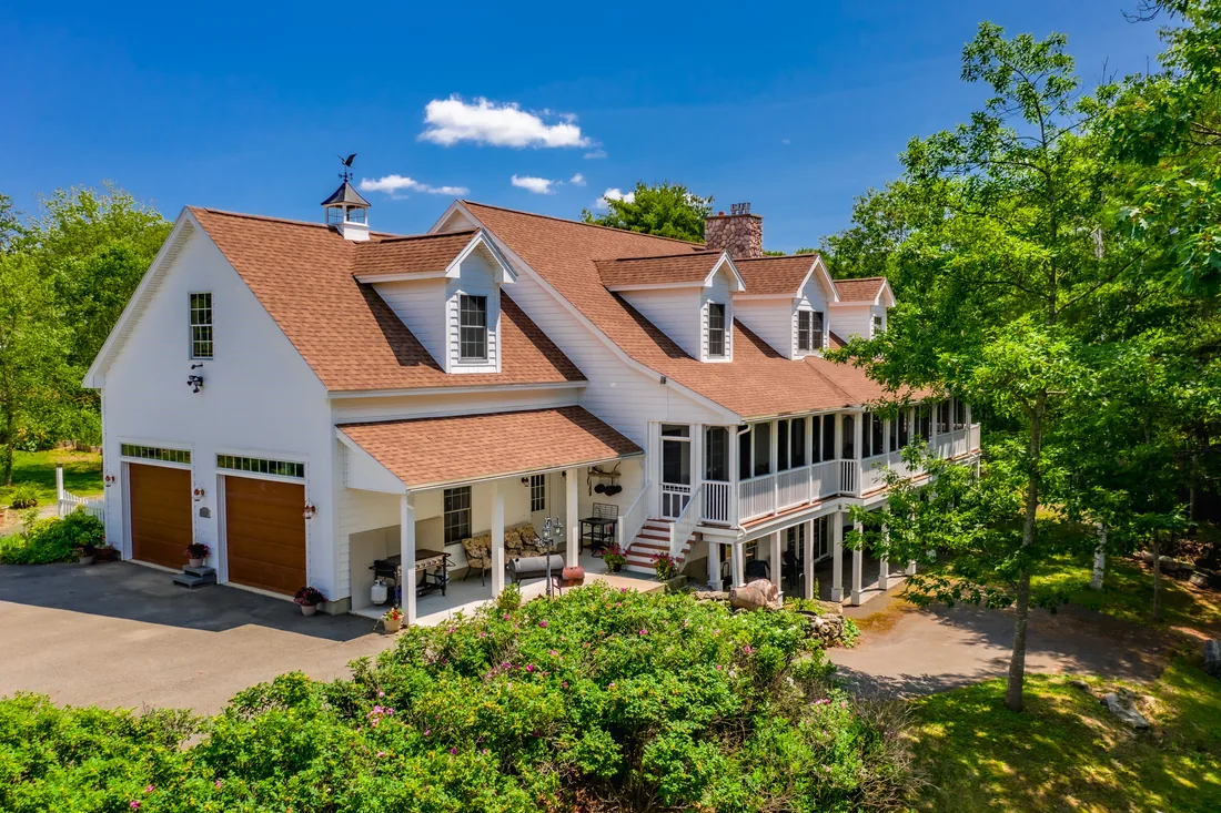 Low aerial view of a large white home with a cupola, a row of dormers, a two-car garage and a long screened porch above an open lower porch, framed by a flowering hedge under a blue summer sky