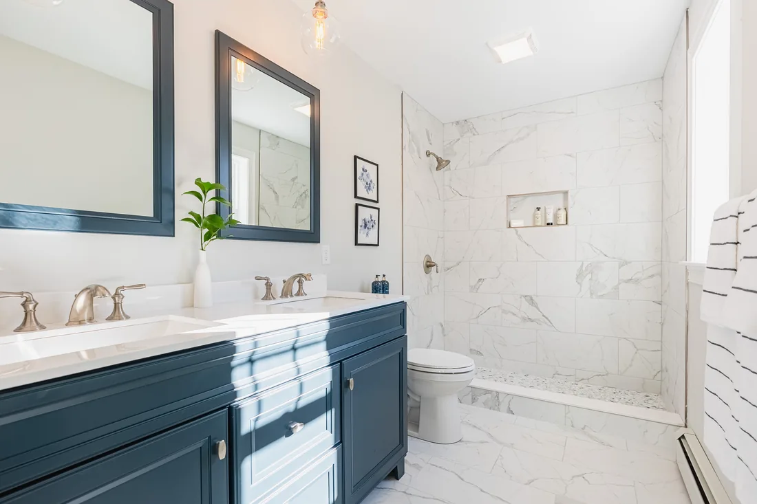 Bathroom with a navy double vanity, brushed nickel faucets and a walk-in shower lined in white marble-look tile