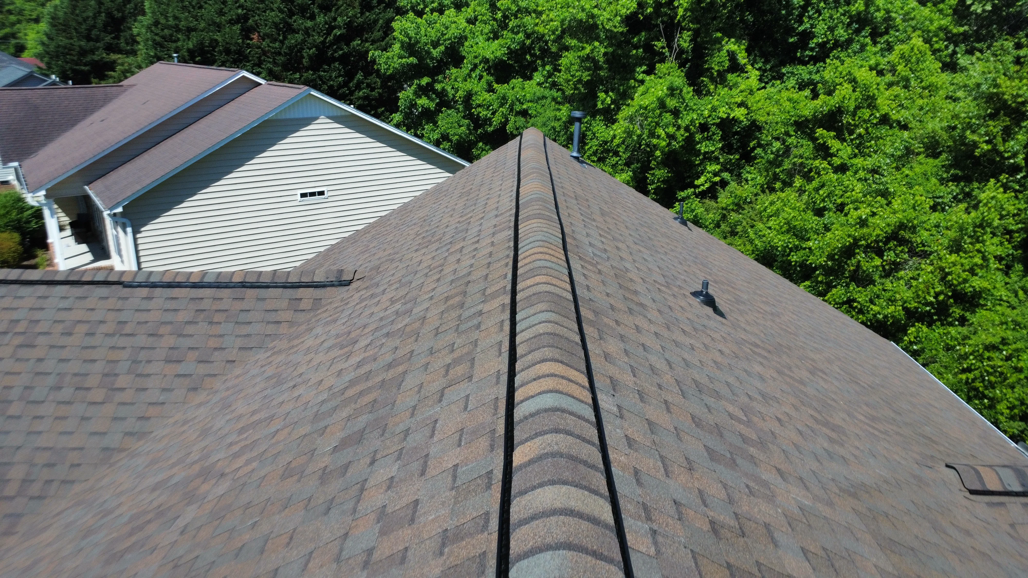 An aerial view of a brown and gray shingle roof with houses and vibrant green trees in the background.