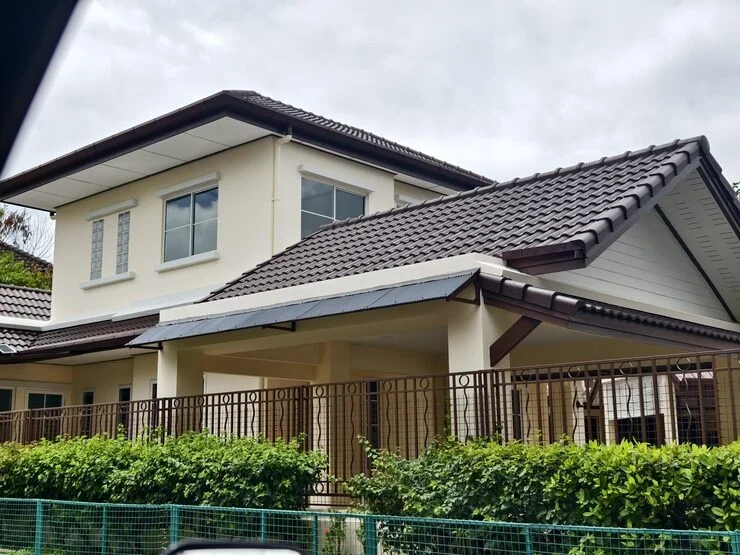 A light-colored two-story house with a brown tiled roof and a brown metal fence in front with green bushes.