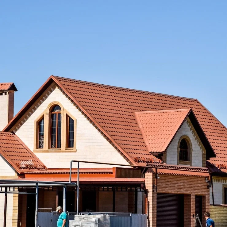 A modern new house with light-colored brick siding, a complex red-brown tiled roof, and a visible garage door. 