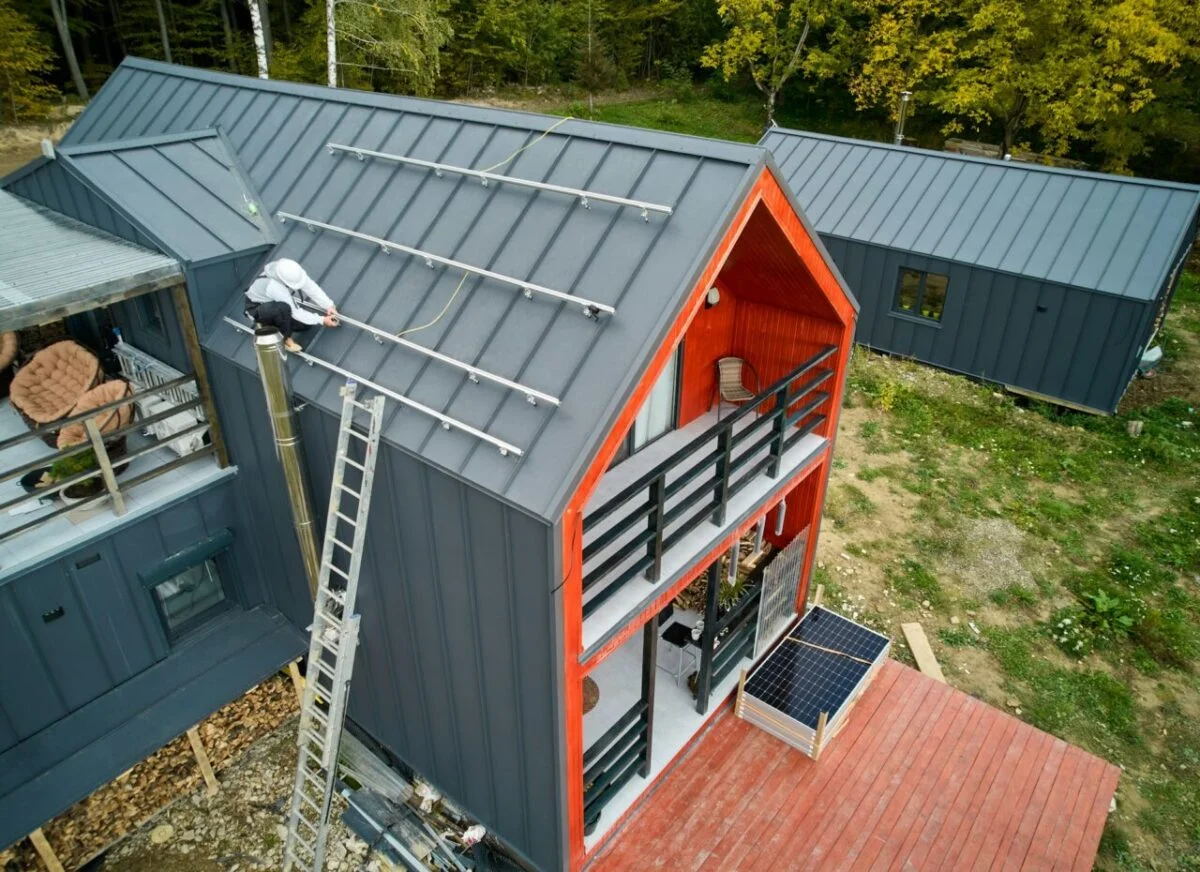 An aerial view of a contractor installing solar panel frames on the dark gray roof of a modern red and gray cabin.